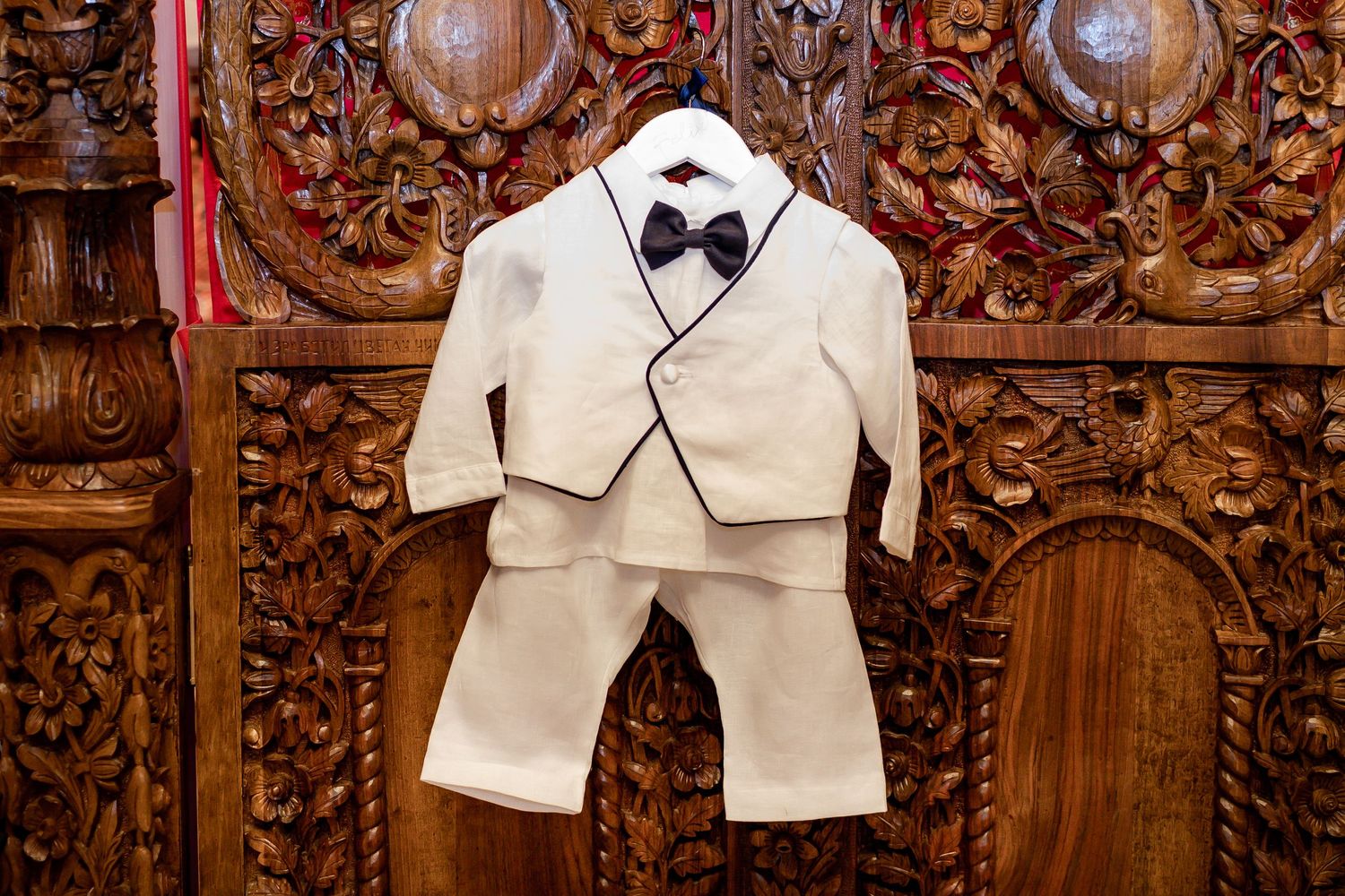 A small white formal suit with black bow tie hangs on display against an ornately carved wooden backdrop.