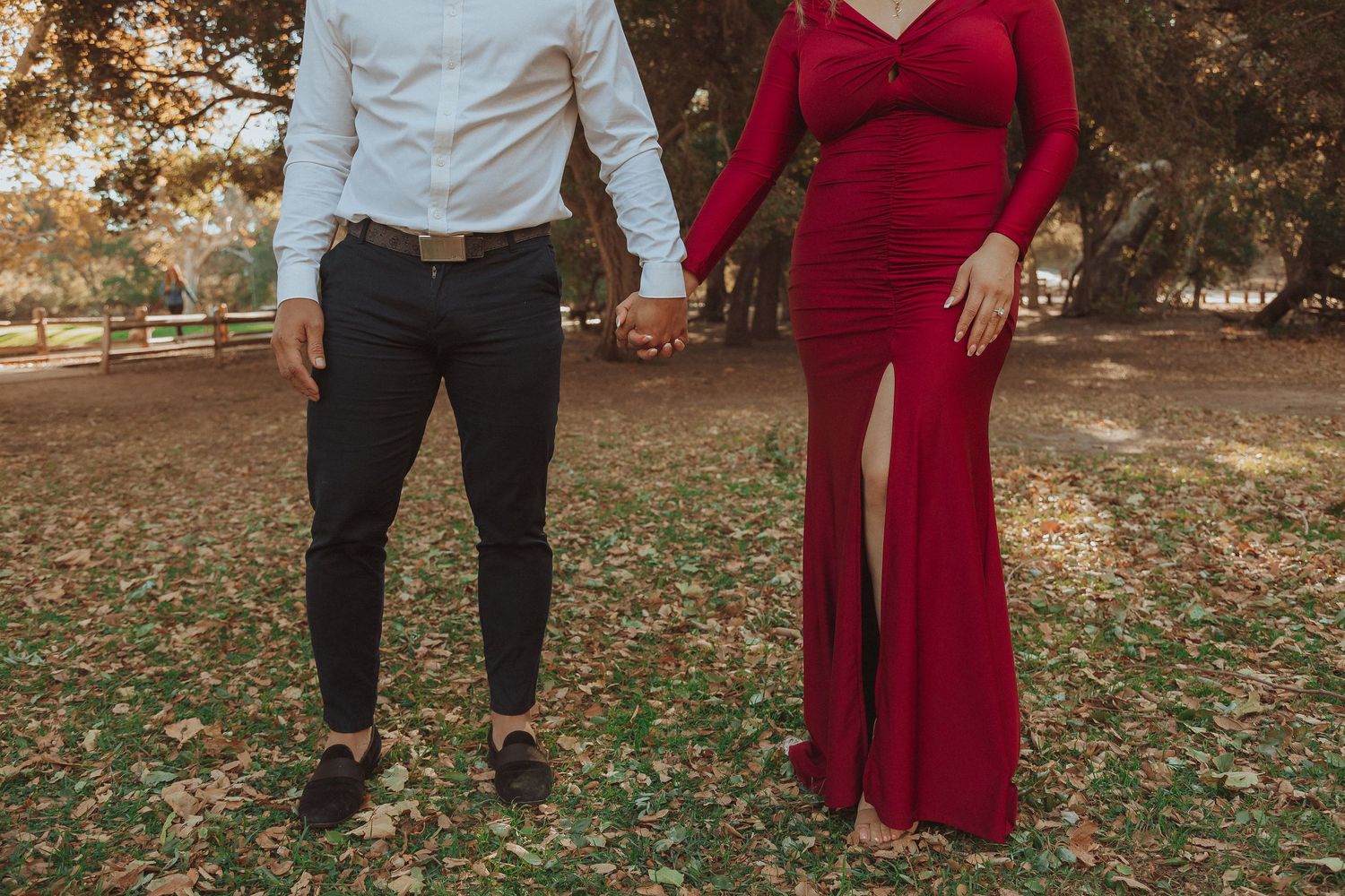Couple in formal attire holding hands while posing in autumn outdoor setting.