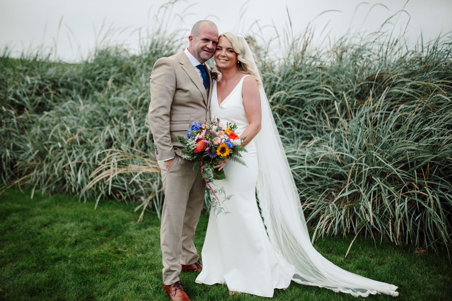 A wedding couple embraces in front of tall ornamental grasses, with the bride holding a vibrant autumn bouquet.