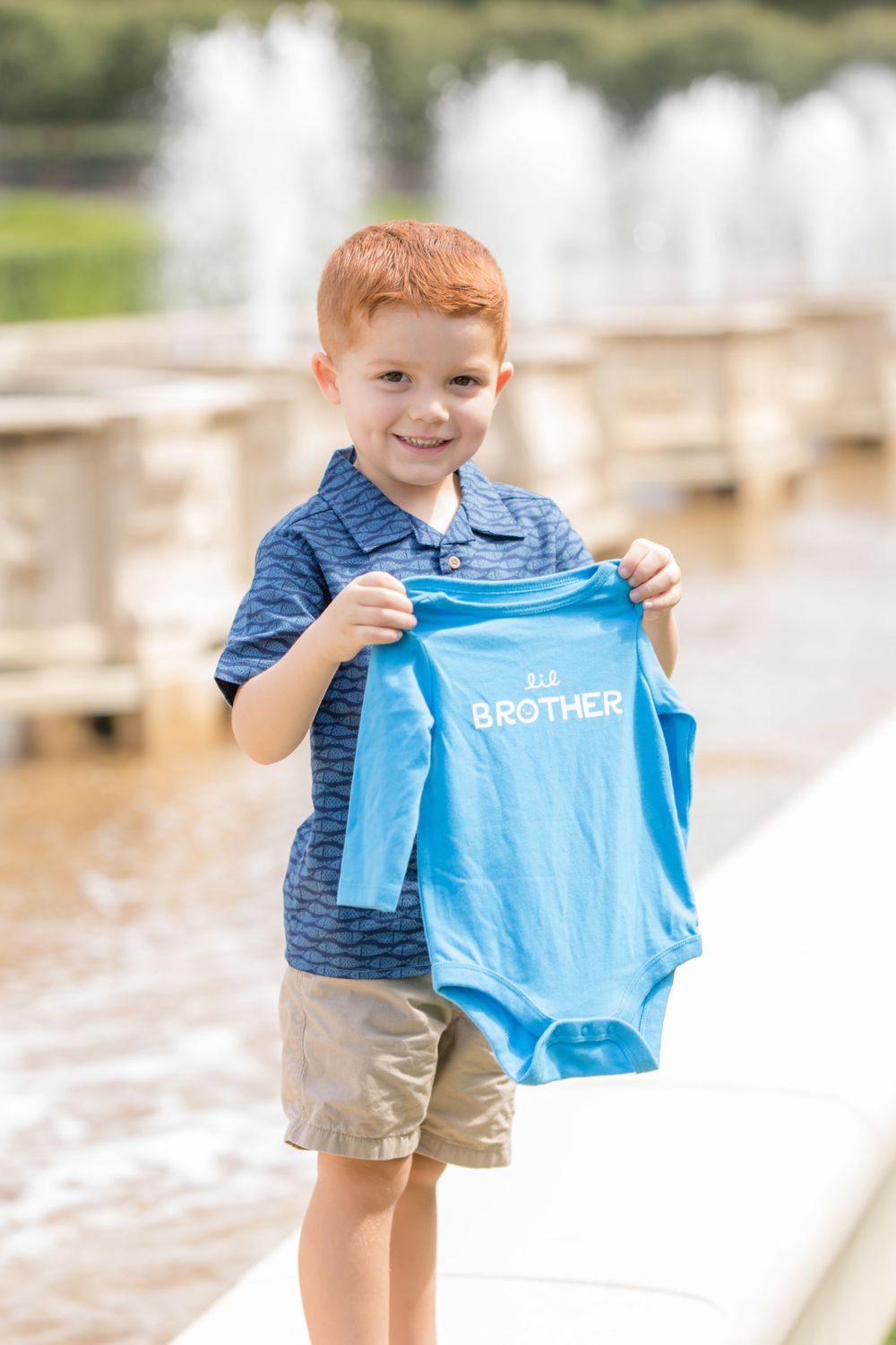 redheaded male child holding onesie in front of limestone fountains for pregnancy announcement in longwood gardens pa