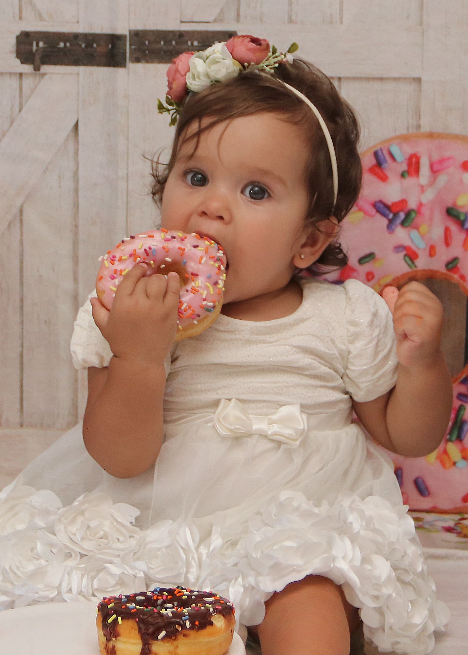 Baby in white dress eating pink frosted donut against white wooden backdrop.