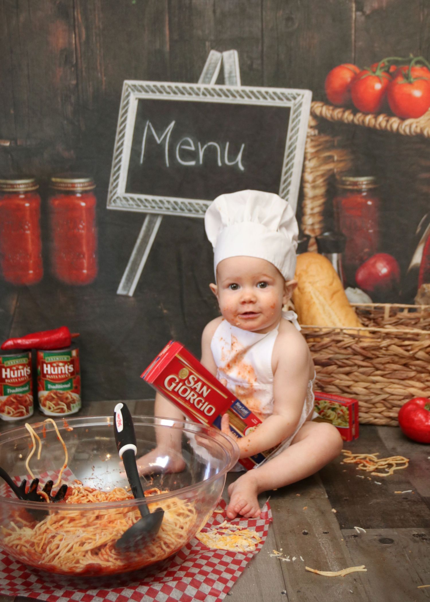 Cooking themed photo setup with pasta, jars of sauce, chef hat, and chalkboard menu sign.