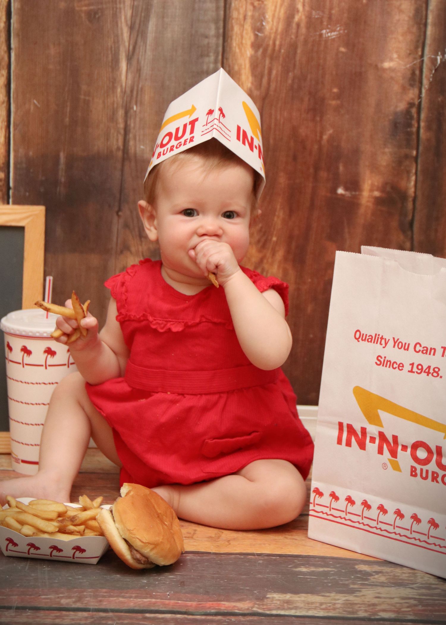 A baby in a red dress enjoys In-N-Out Burger with fries and a drink while wearing a paper hat.