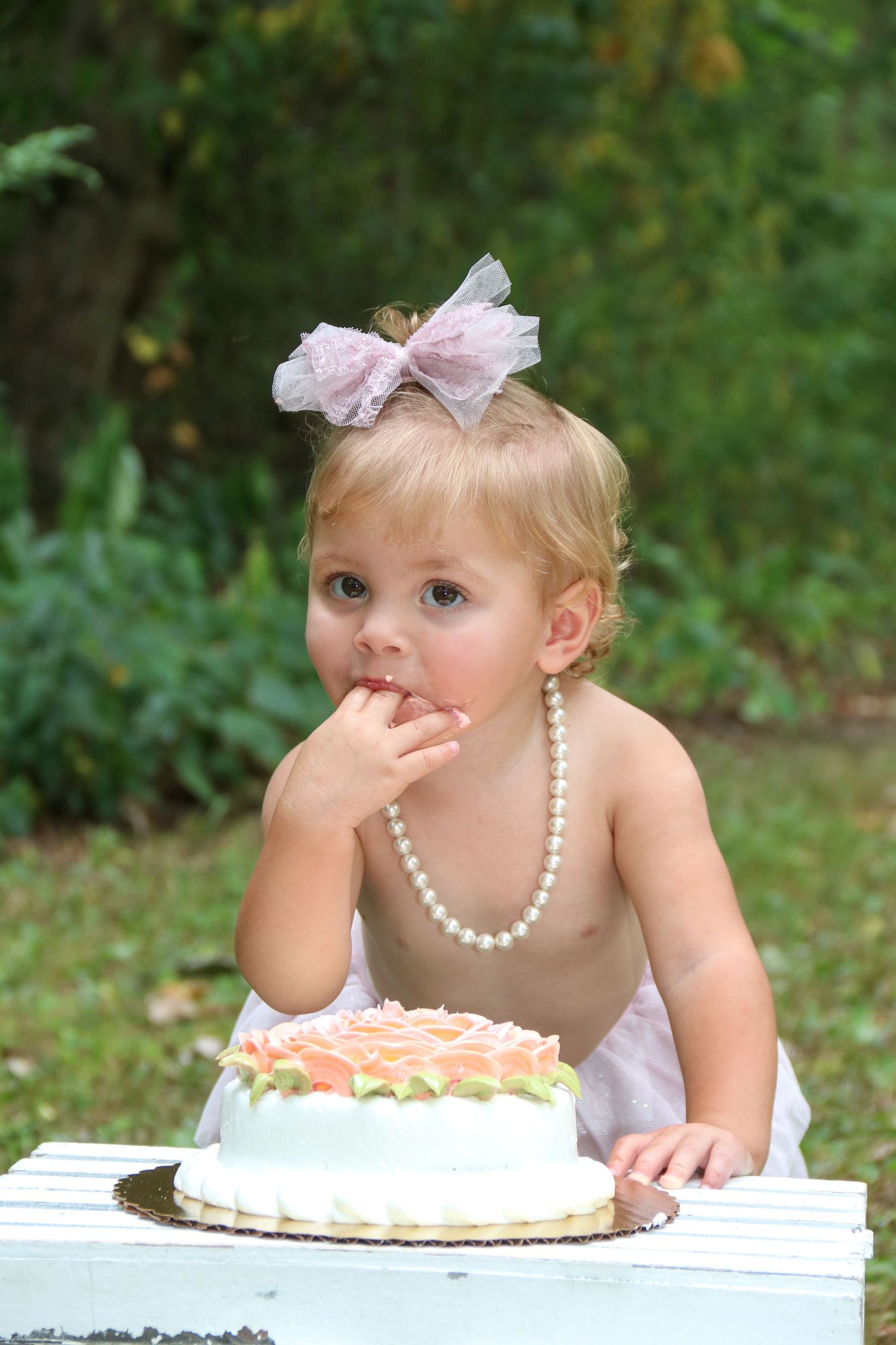 A cake smash photo setup with a white cake on a wooden surface and pearl necklace accessory.