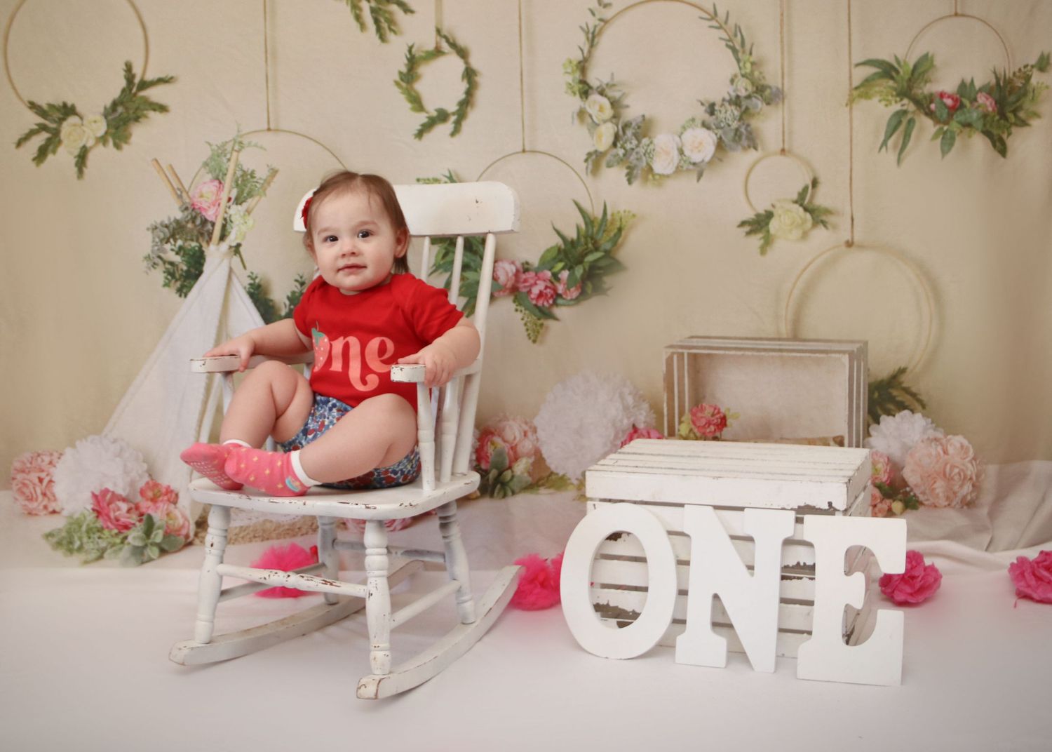 First birthday photo setup with white rocking chair and floral wall decorations.