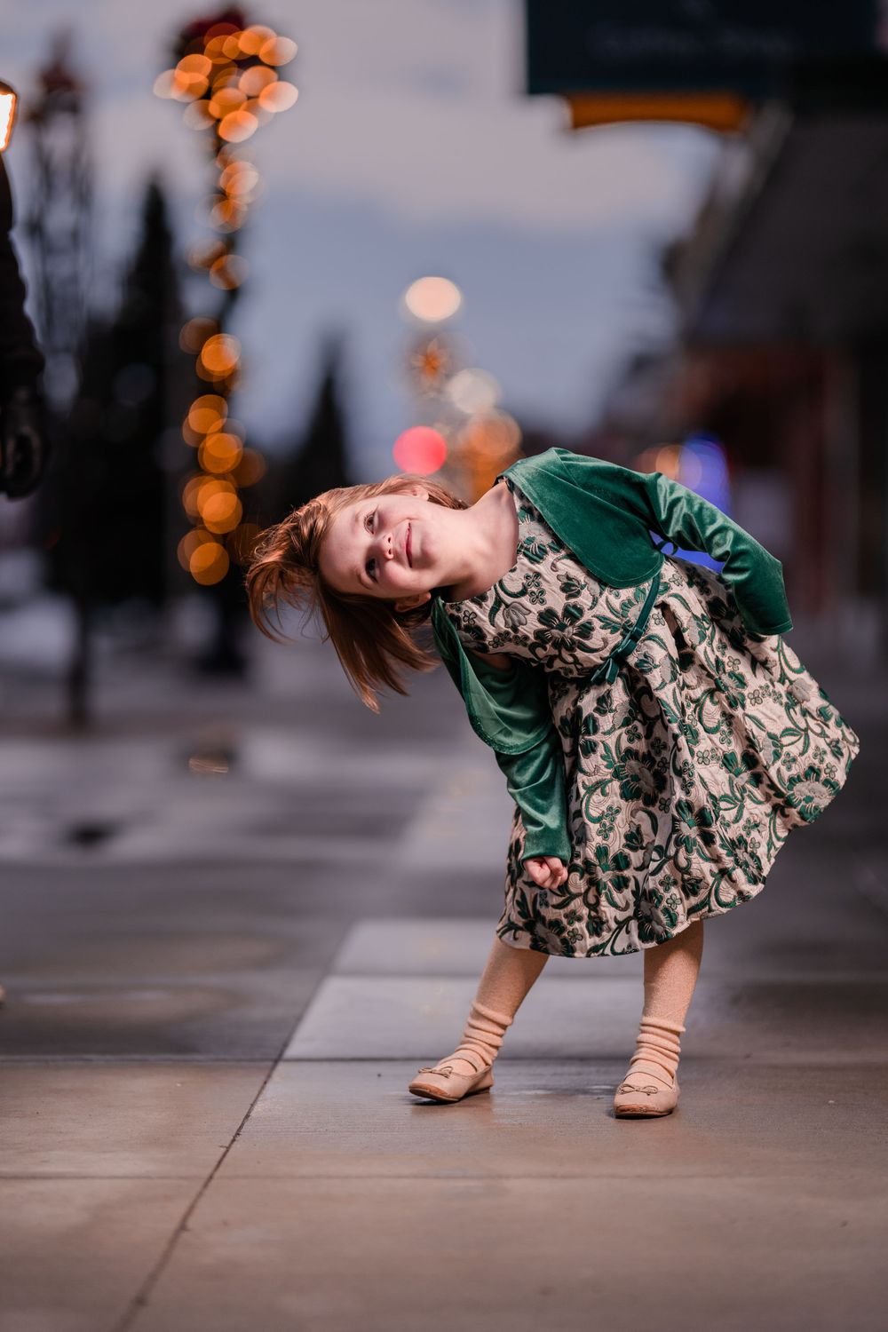 Child in green dress playfully poses on city sidewalk with bokeh lights in background.