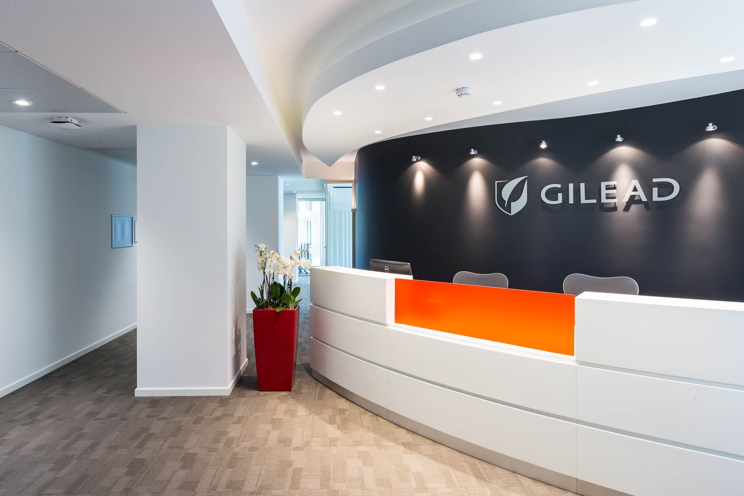 Modern office reception area with white curved desk and Gilead logo on dark wall with orange accent panels.