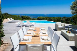 Luxury outdoor dining setup with ocean view, infinity pool and white furniture on wooden deck.