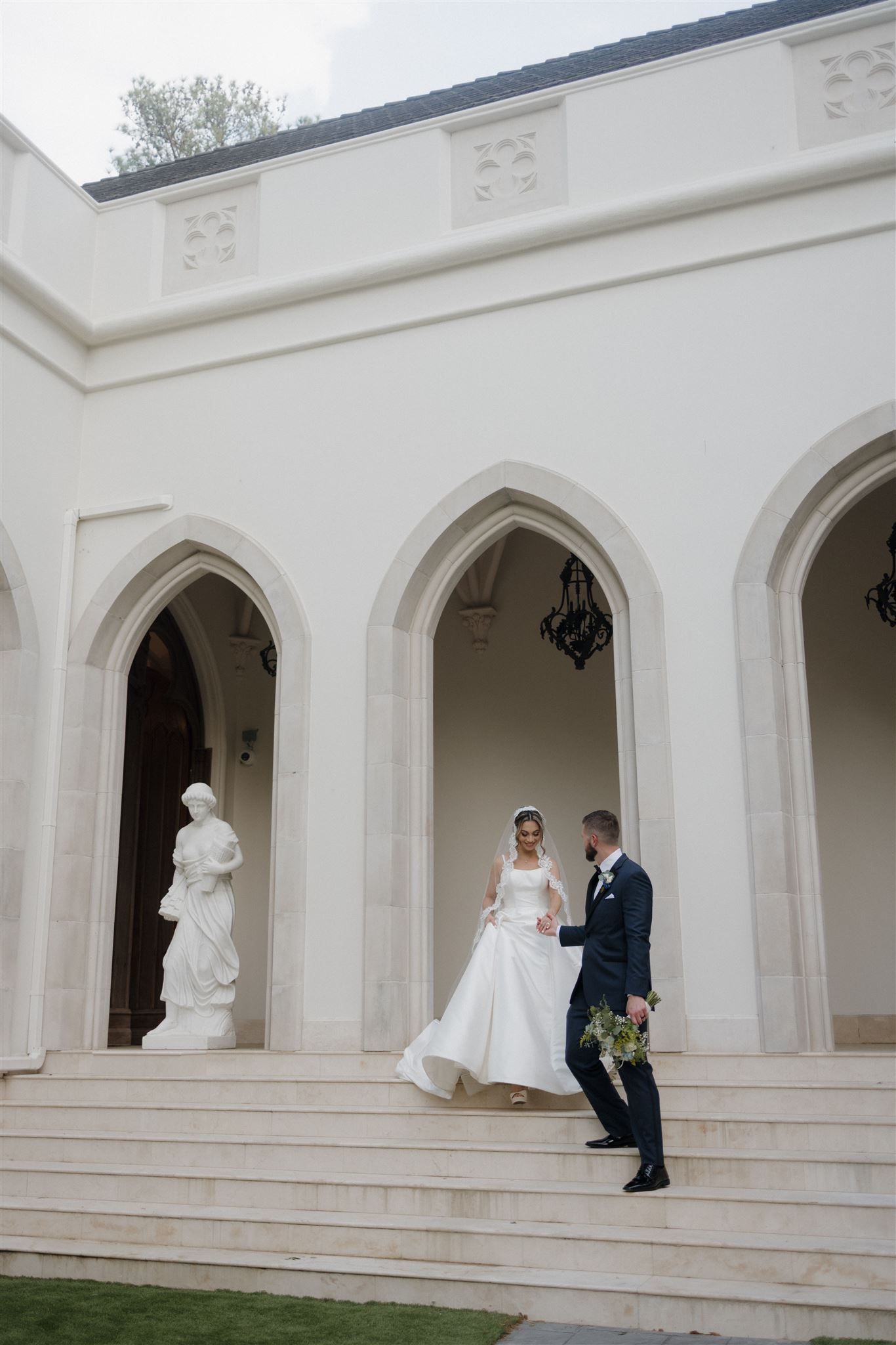 Couple stands on steps of white gothic-style building with arched alcoves and classical statues.