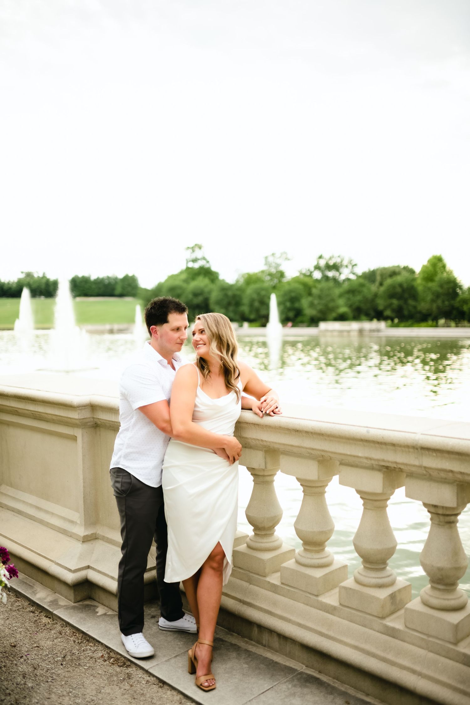 A romantic couple embracing on a stone balcony overlooking a lake with fountains.