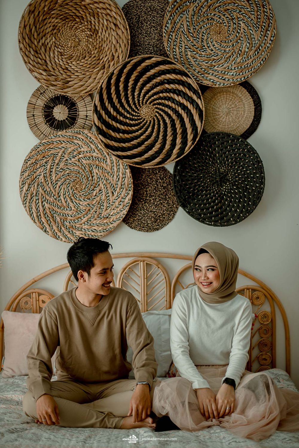 Couple sitting on bed with decorative woven circular wall art above in neutral tones.