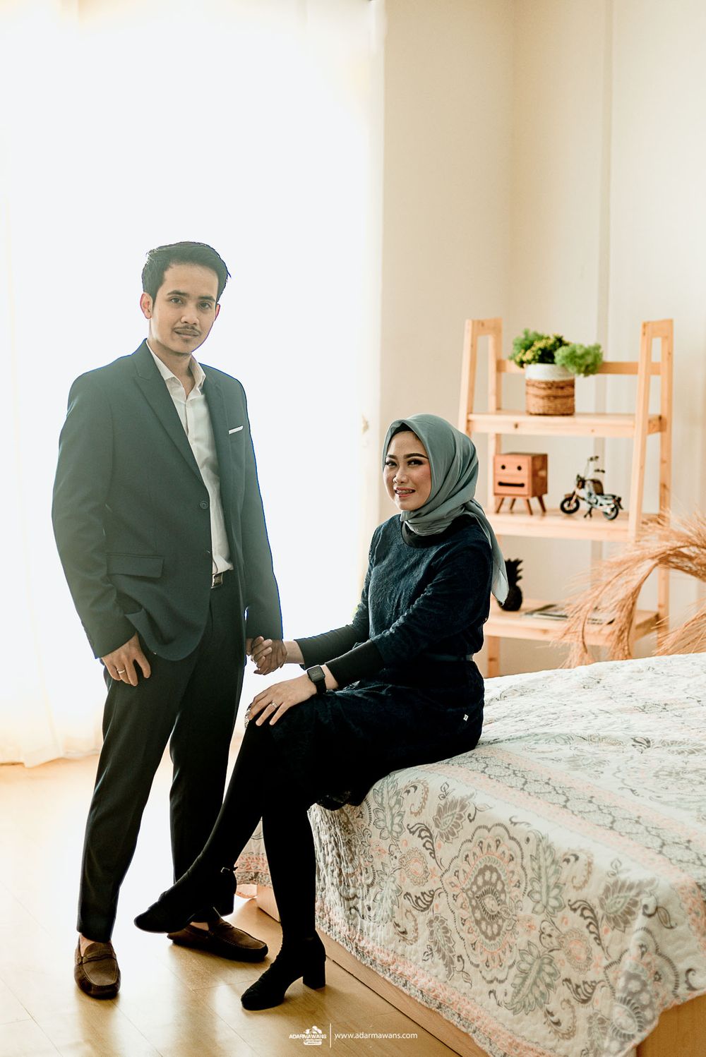 Elegant couple poses in a cozy home setting with modern decor and a bed in the background.