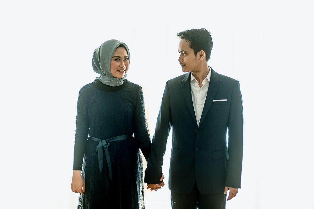 Couple in matching formal black attire holding hands while standing against white background.