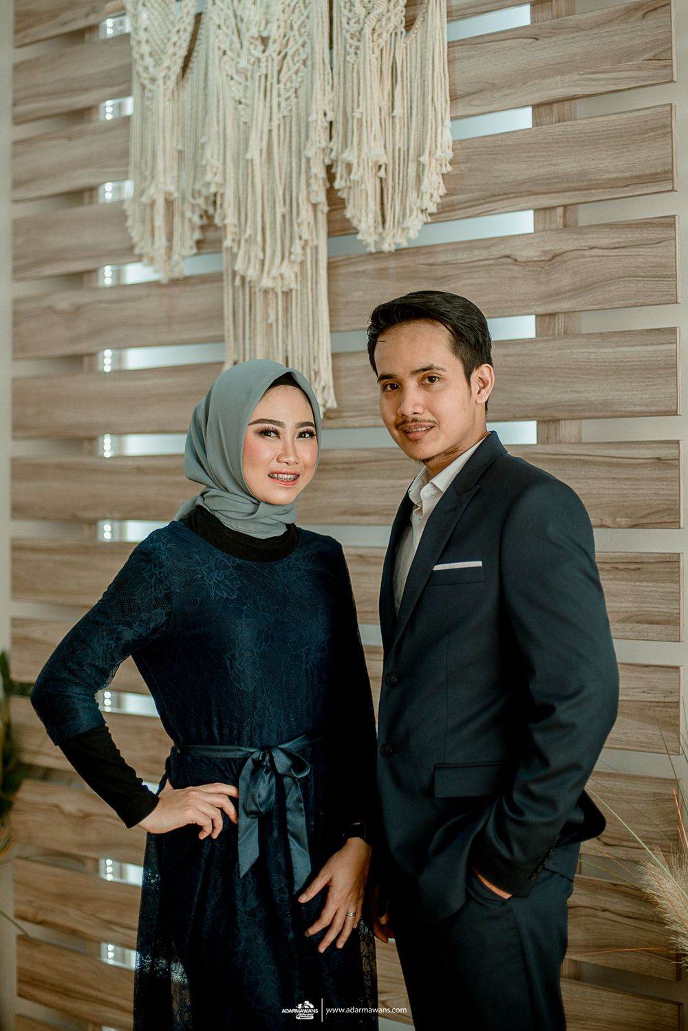 Elegant couple poses in formal attire against decorative macrame wall hanging.