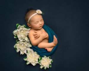 Newborn photography with baby wrapped in teal fabric surrounded by white lotuses against dark blue background.
