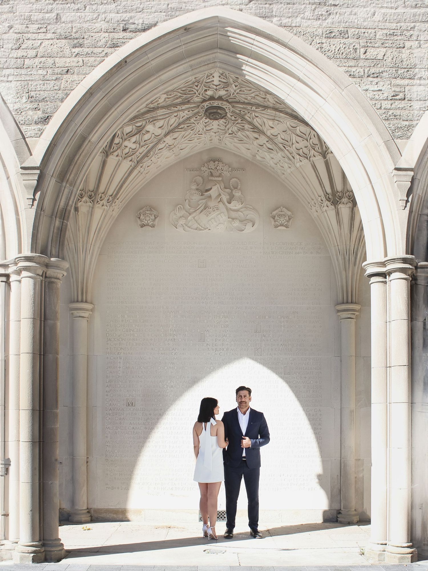 A couple stands together under an ornate white stone archway with gothic architectural details.