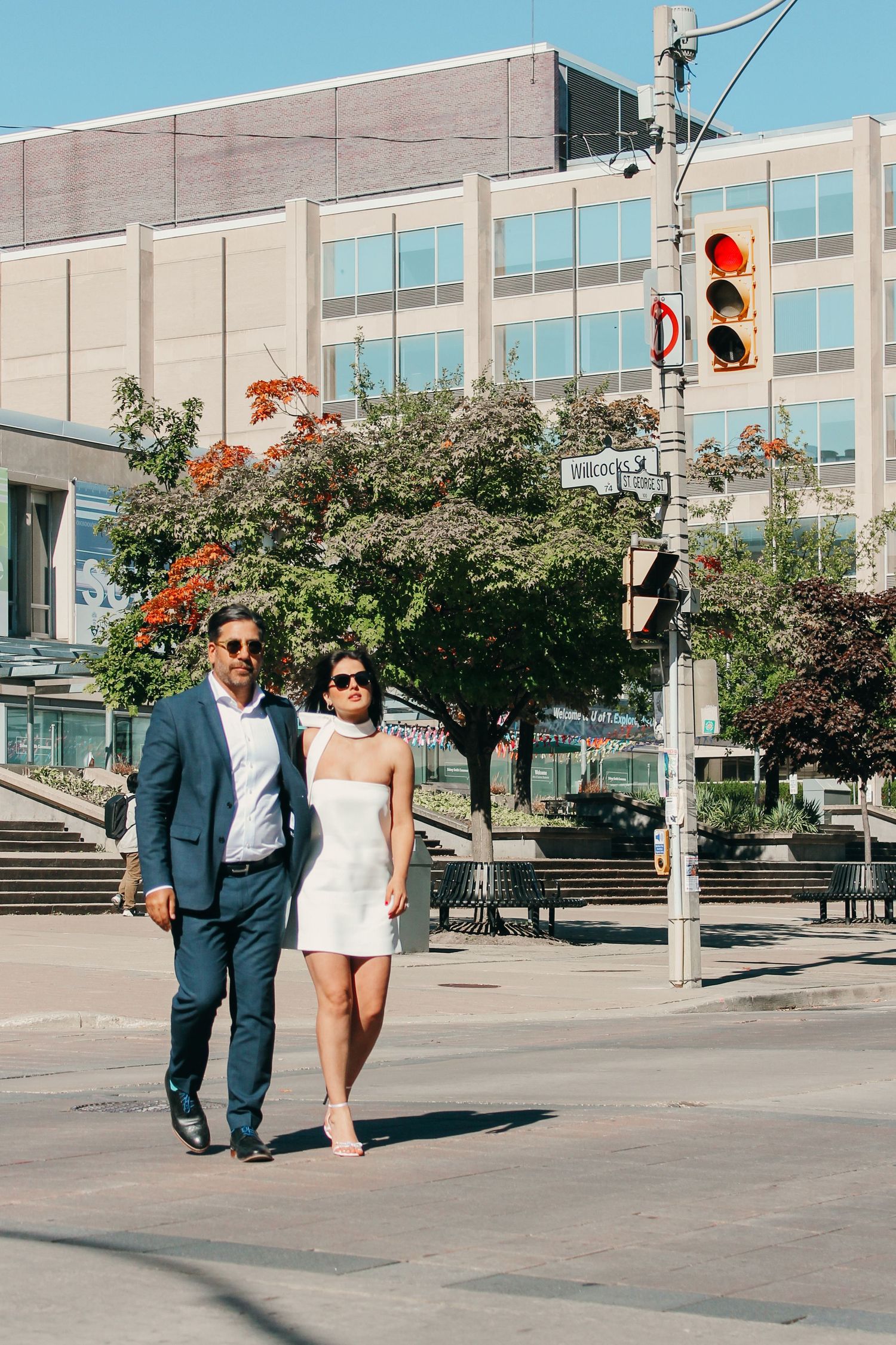 A couple in formal attire walking together on a city street with traffic lights and buildings in the background.