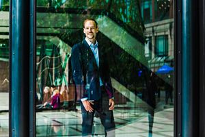 Executive in navy suit poses confidently against reflective glass building with urban greenery.