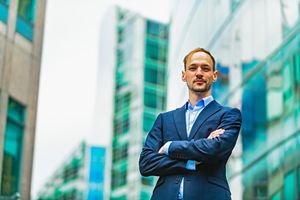 Professional businessman standing confidently with arms crossed outside modern office buildings in urban setting.