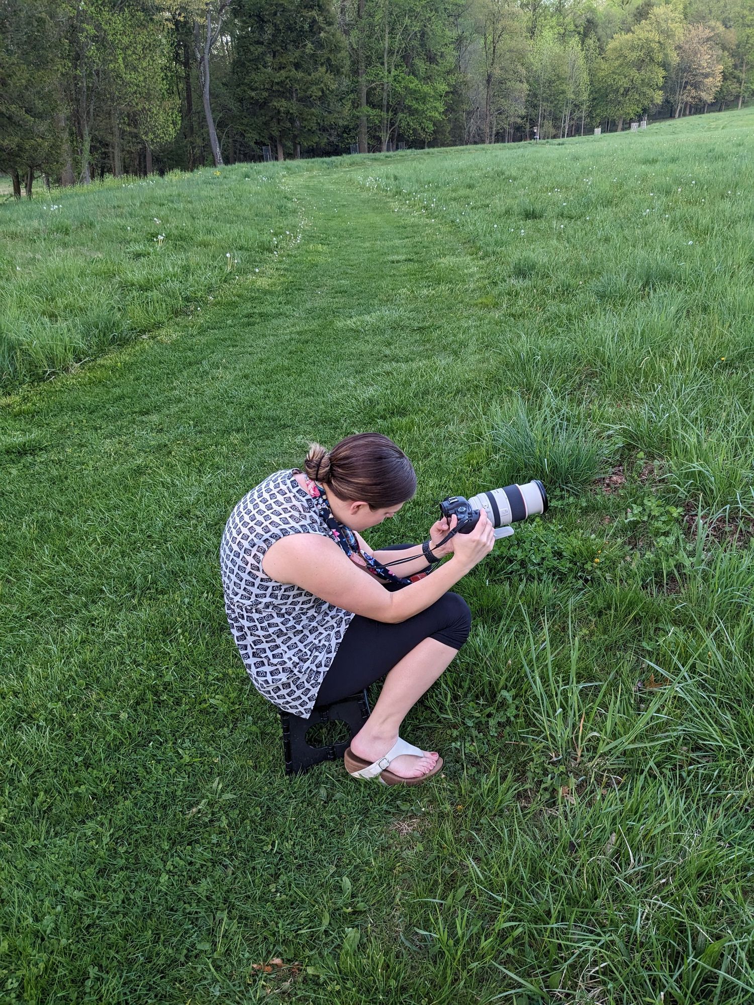 Photographer in patterned dress crouches with camera in grassy field.