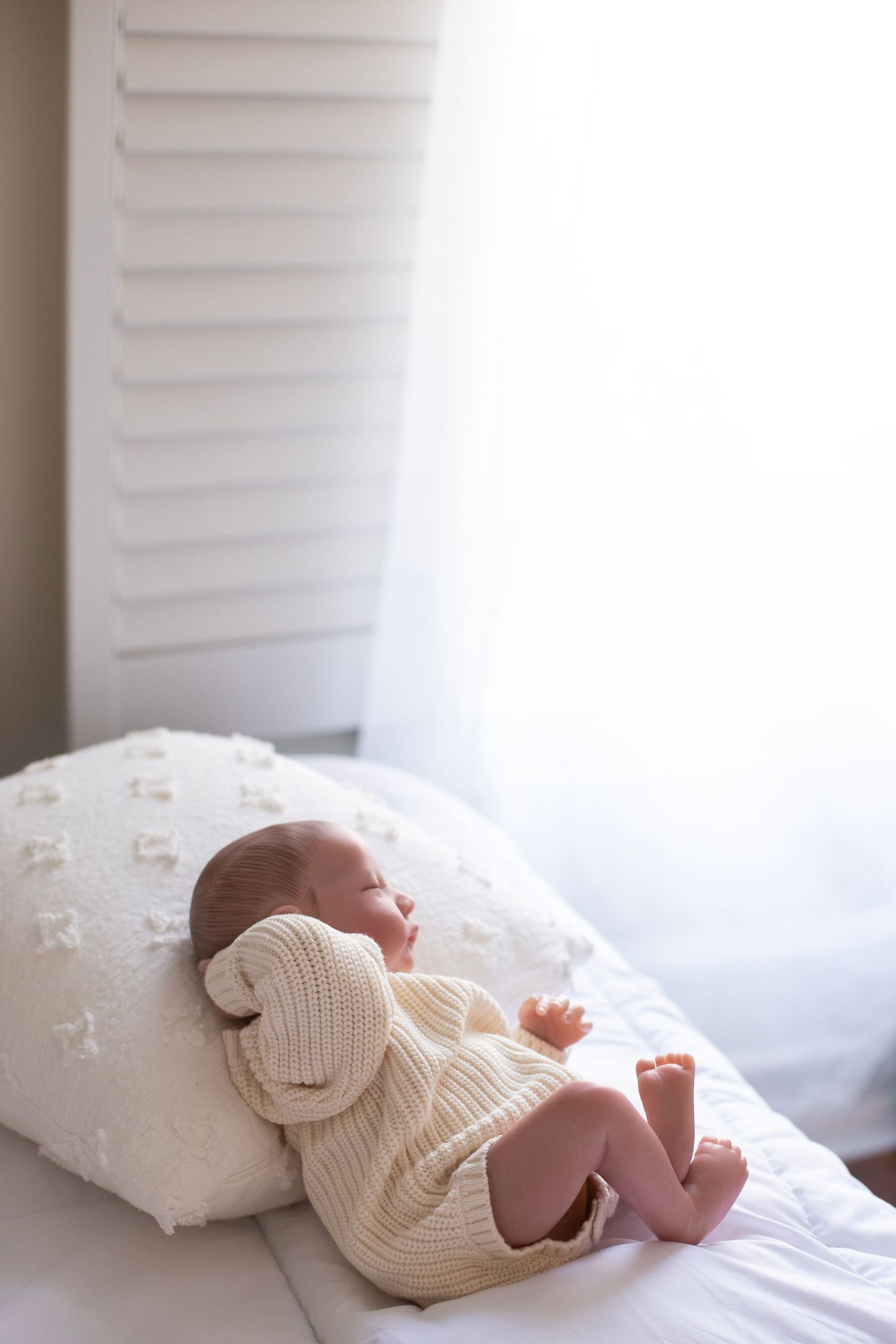 Baby doll laying on a bed, near a window, wearing a sweater with it's head resting on a white pillow.