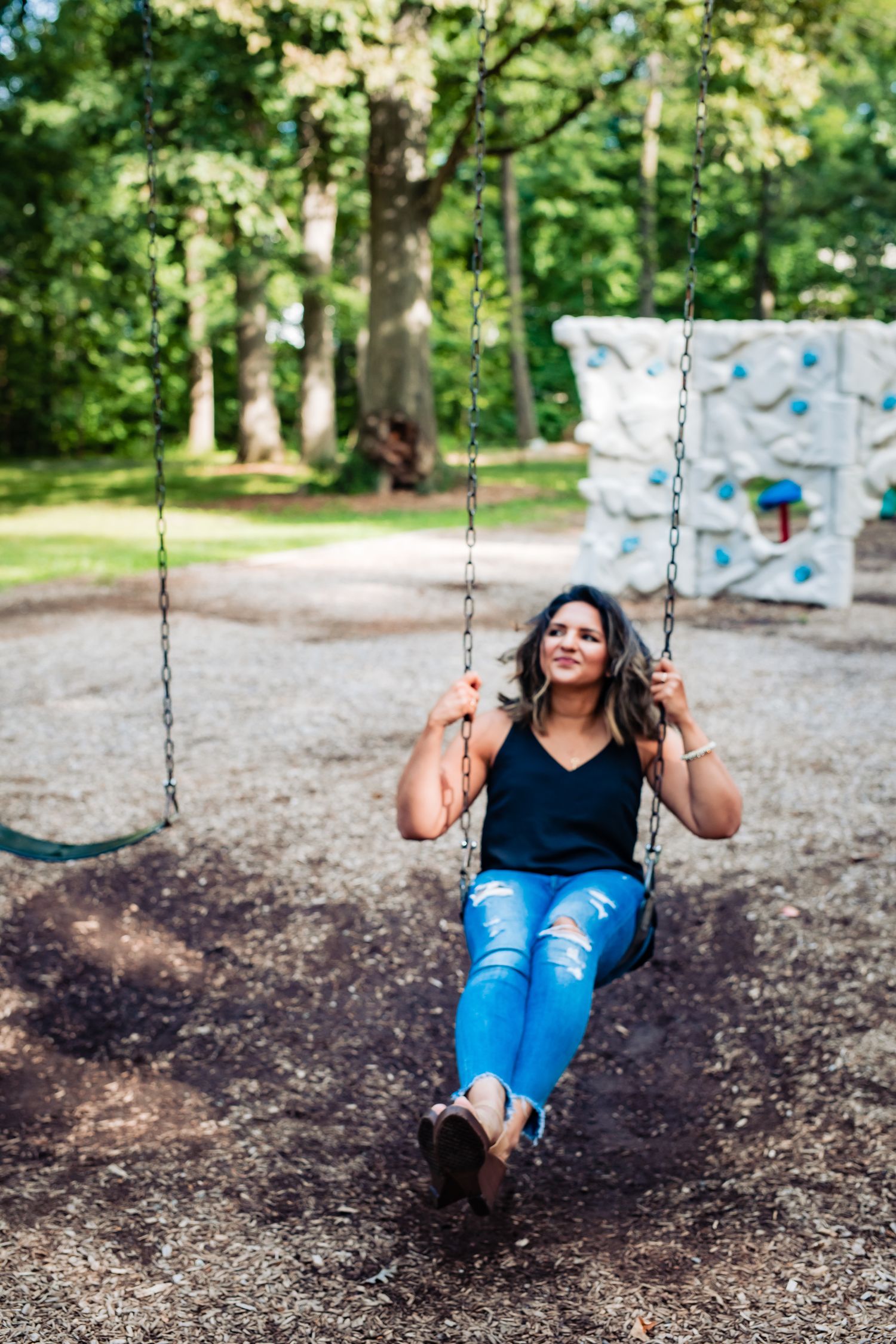Person enjoying swing in park wearing blue jeans and black top in summer setting.