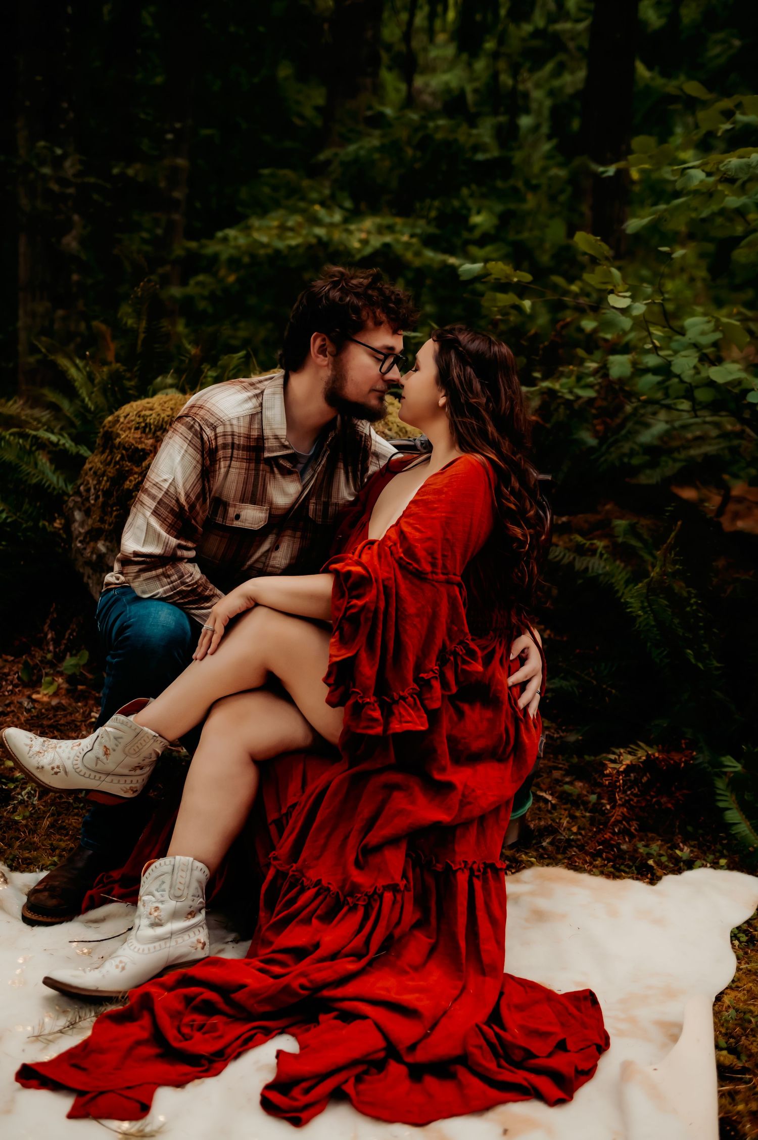 Romantic couple in red dress and plaid shirt posing on snowy ground in forest setting.