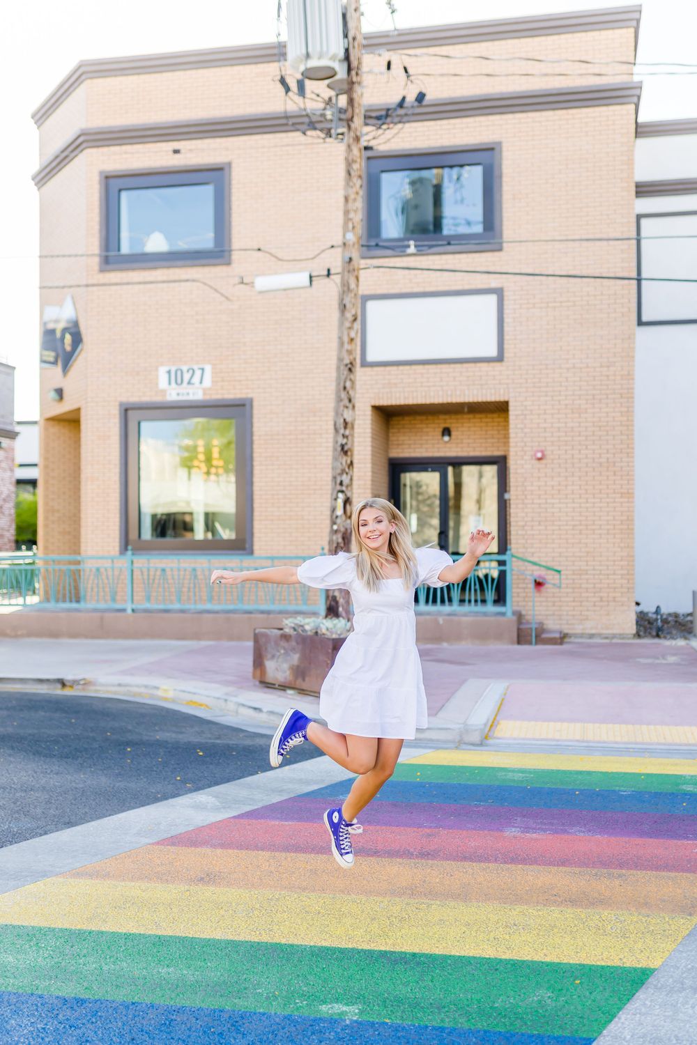 Person jumping joyfully on rainbow crosswalk in front of brick building.