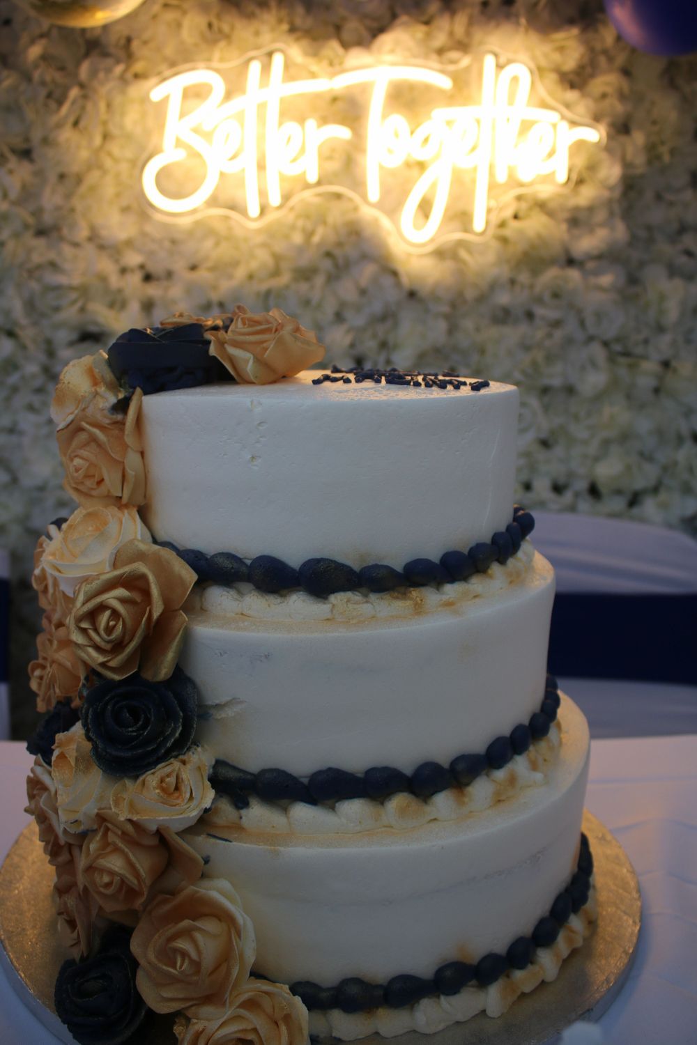 A white wedding cake with navy blue trim and roses sits beneath glowing neon signage.
