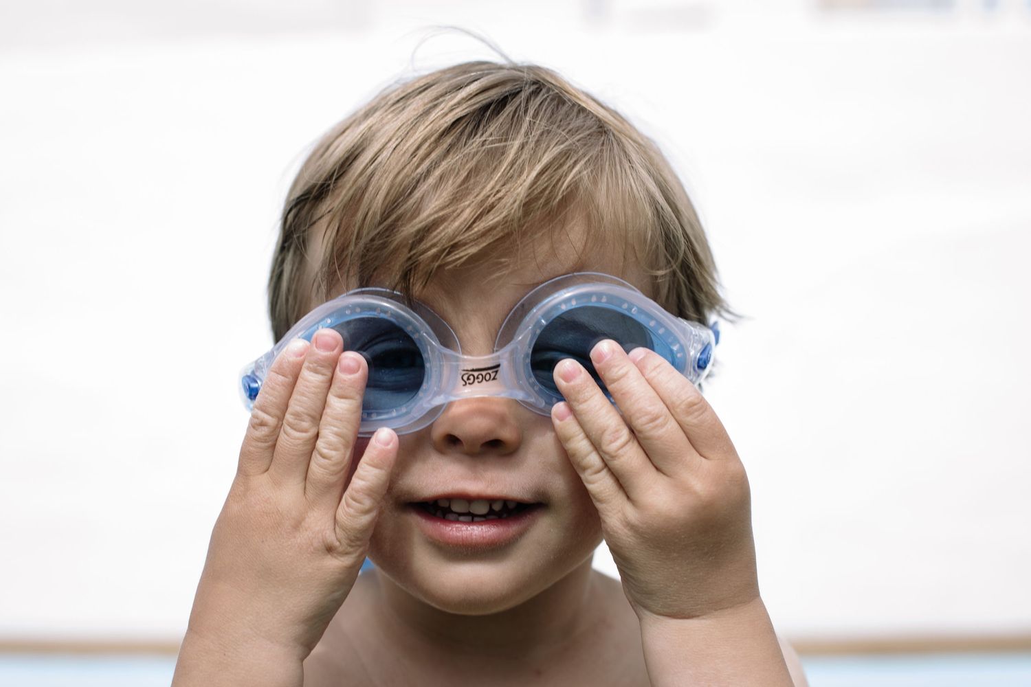 Child wearing blue swimming goggles smiles while holding them with both hands.