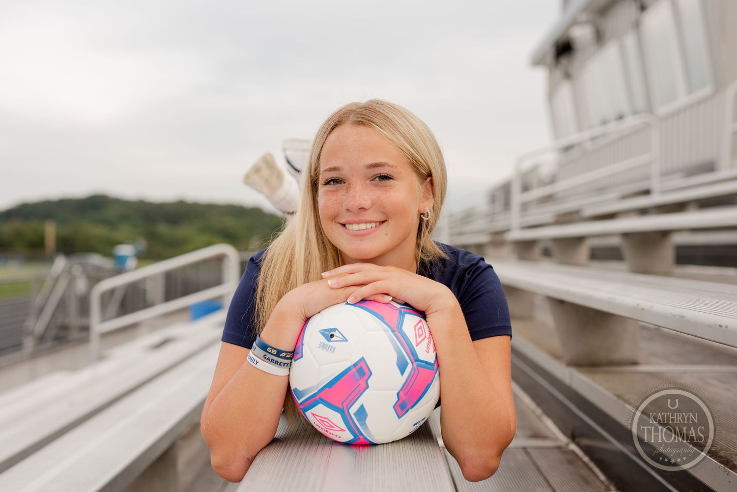 Athletic portrait on bleachers with soccer ball and stadium backdrop during sunny day.