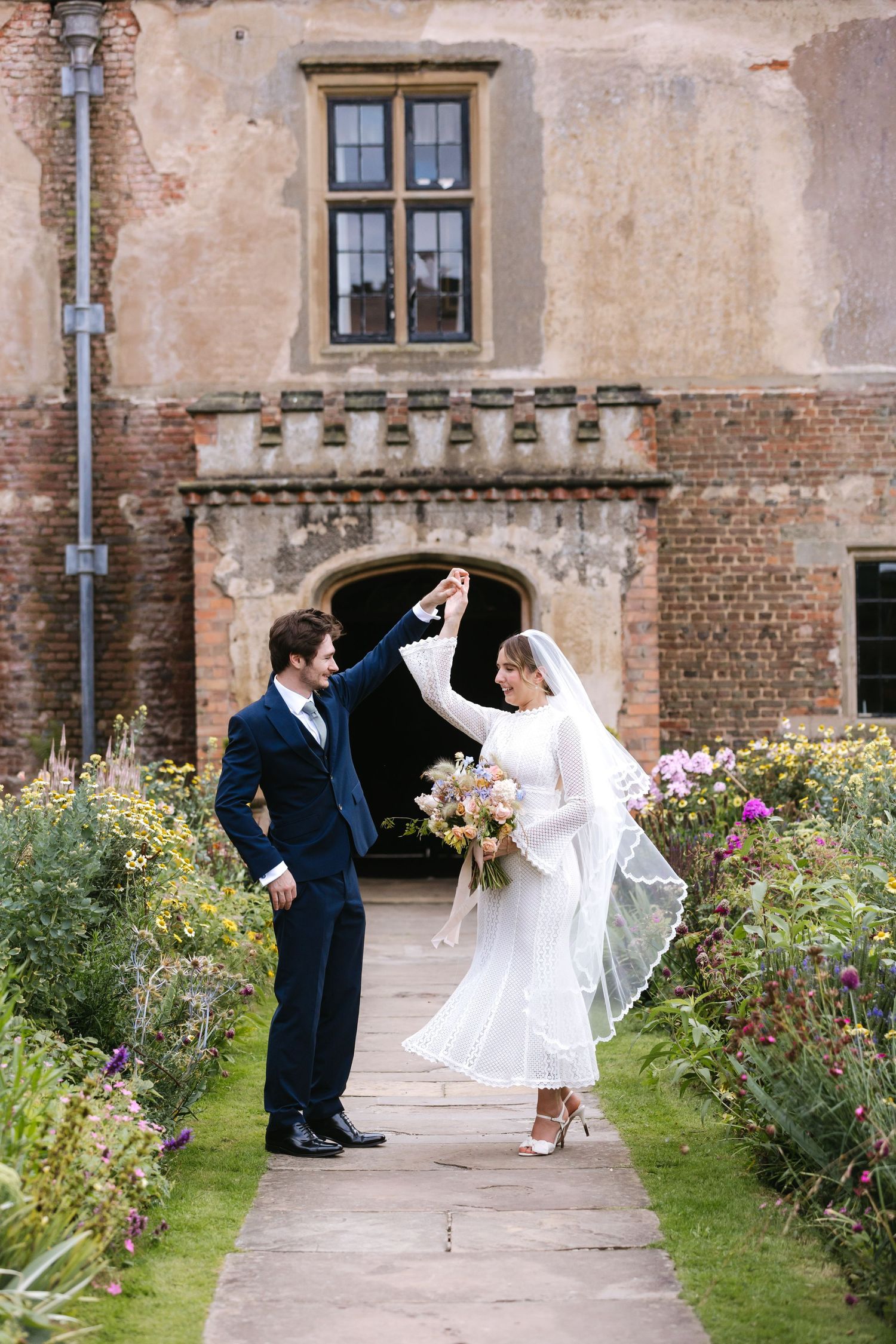 Elegant wedding couple dancing on garden path surrounded by blooming flowers and historic architecture.