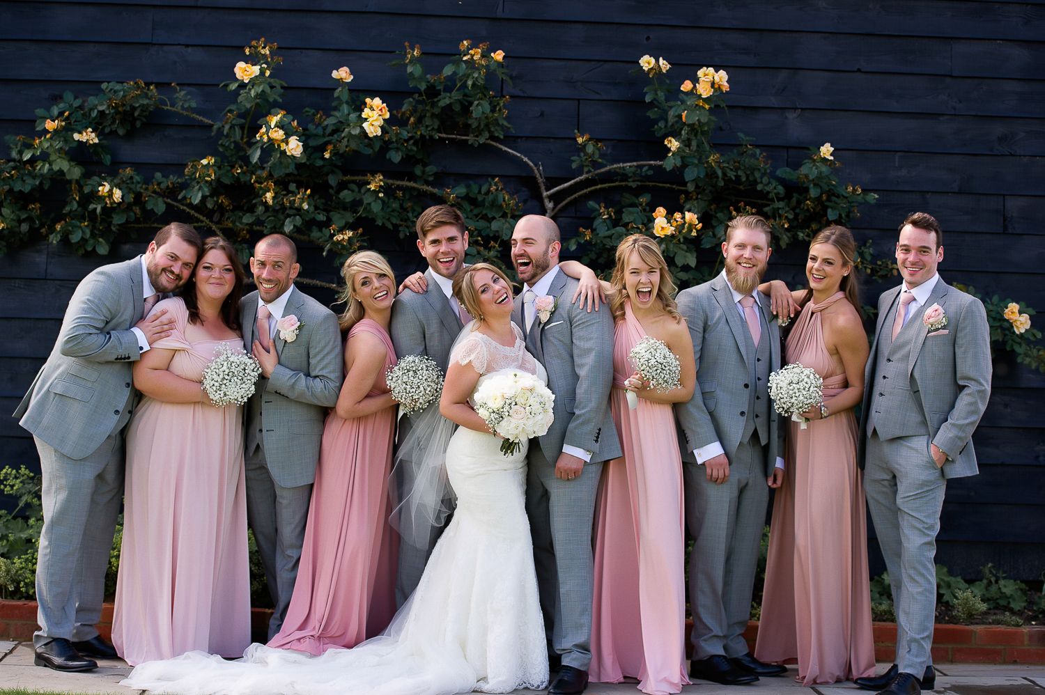 Wedding party in pink dresses and grey suits pose against dark blue wall with climbing roses.