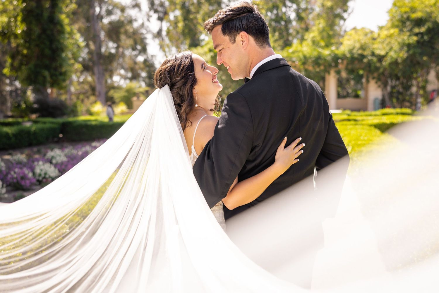 Wedding couple shares intimate moment with bride's veil flowing dramatically in garden setting.