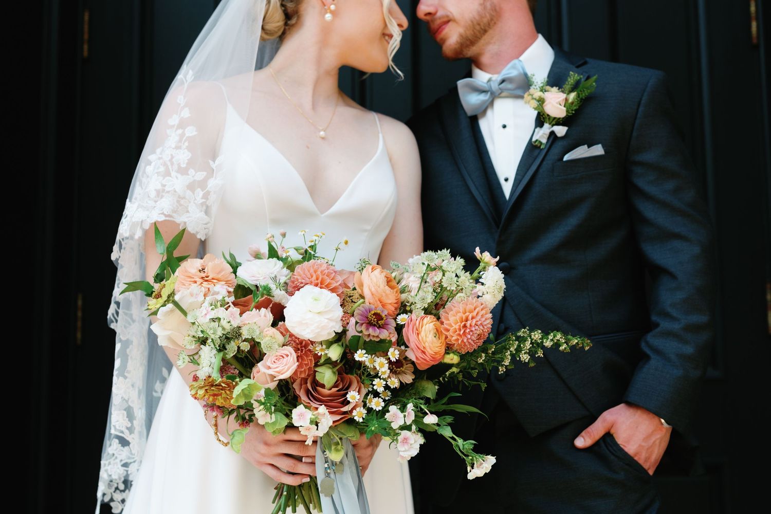 Elegant couple in formal wedding attire with lush peach and white floral bouquet.