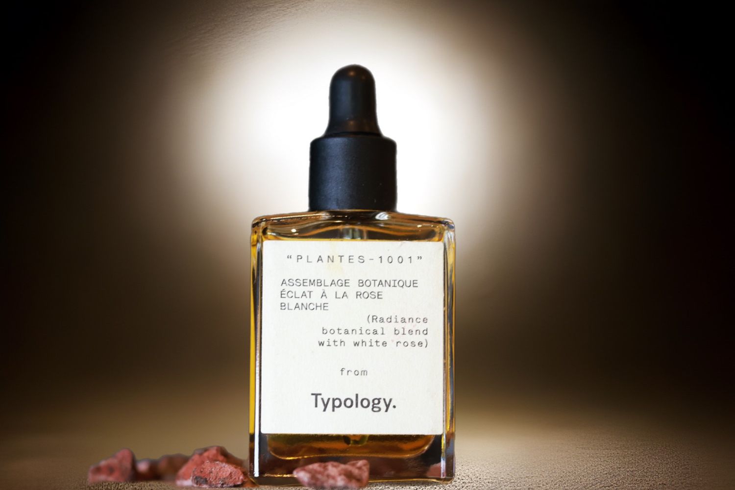 A bottle of Typology skincare serum with dropper against dark background with soft lighting and pink crystals.
