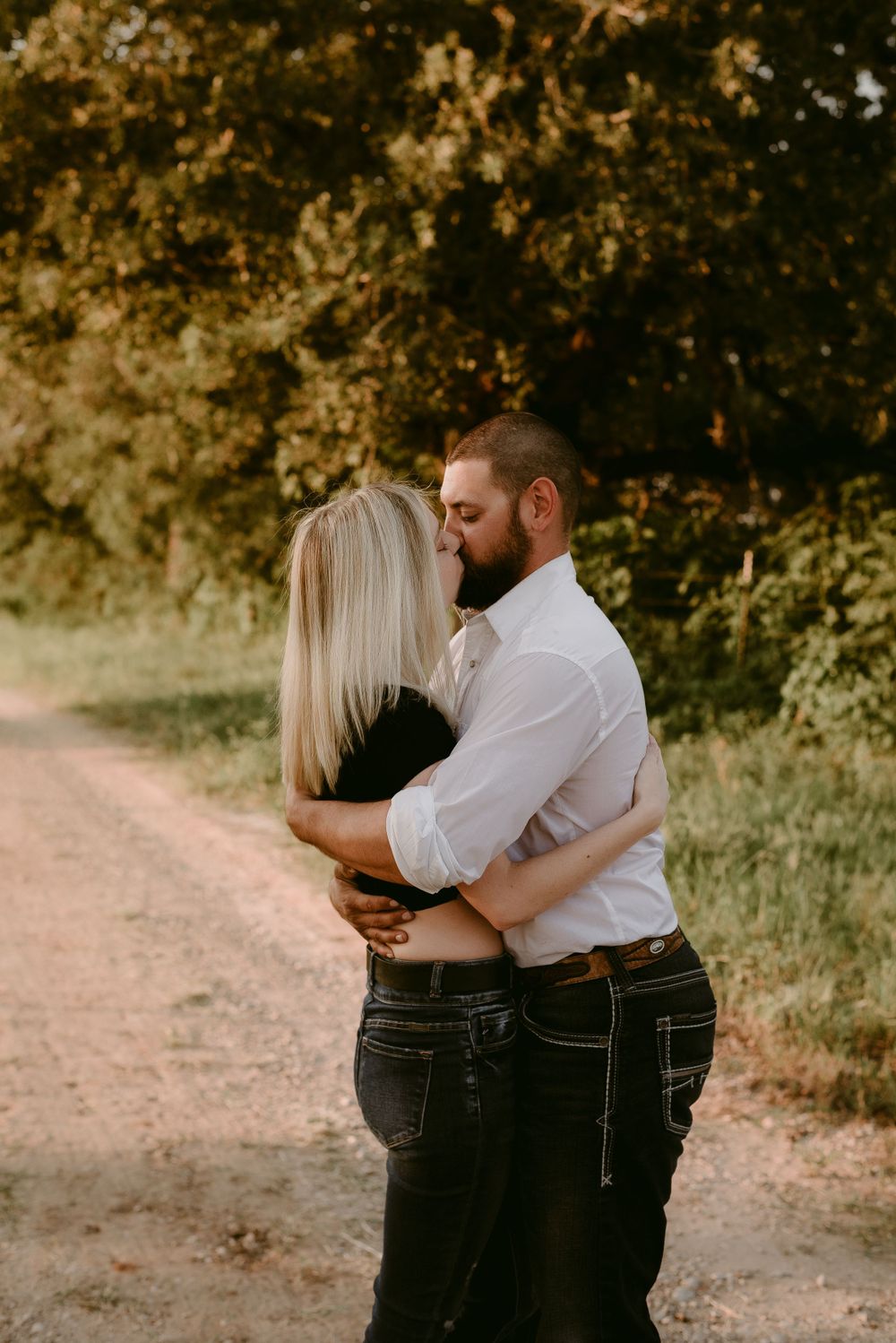 A couple shares an intimate embrace on a sunlit country road with trees in the background.