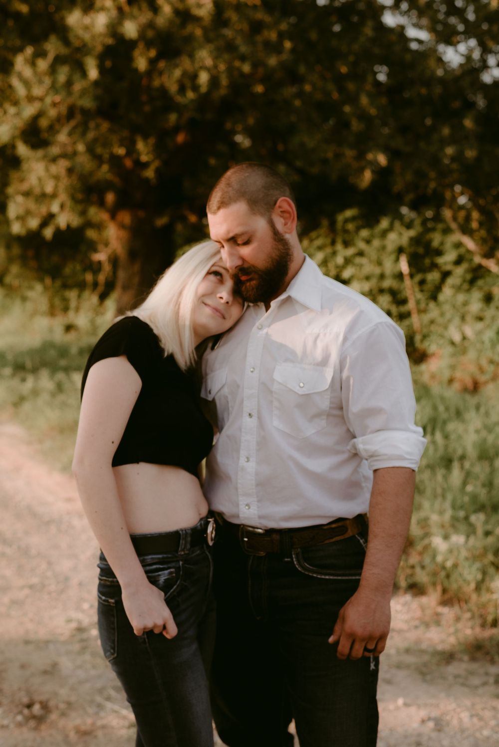 A romantic couple embraces on a dirt path with golden sunlit trees in the background.