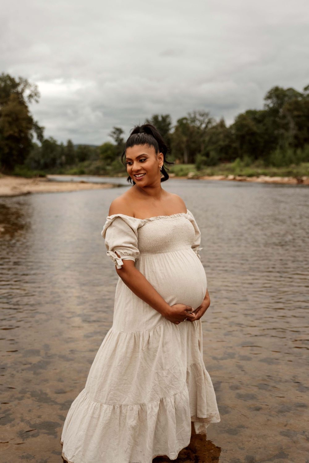 Pregnant woman in white off-shoulder dress standing in shallow river smiling.