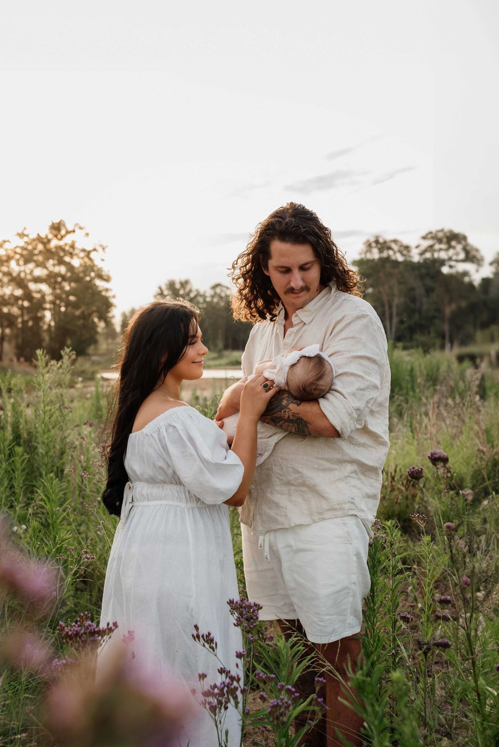 Couple in white outfits admiring newborn baby in wildflower meadow during golden hour sunset shoot.