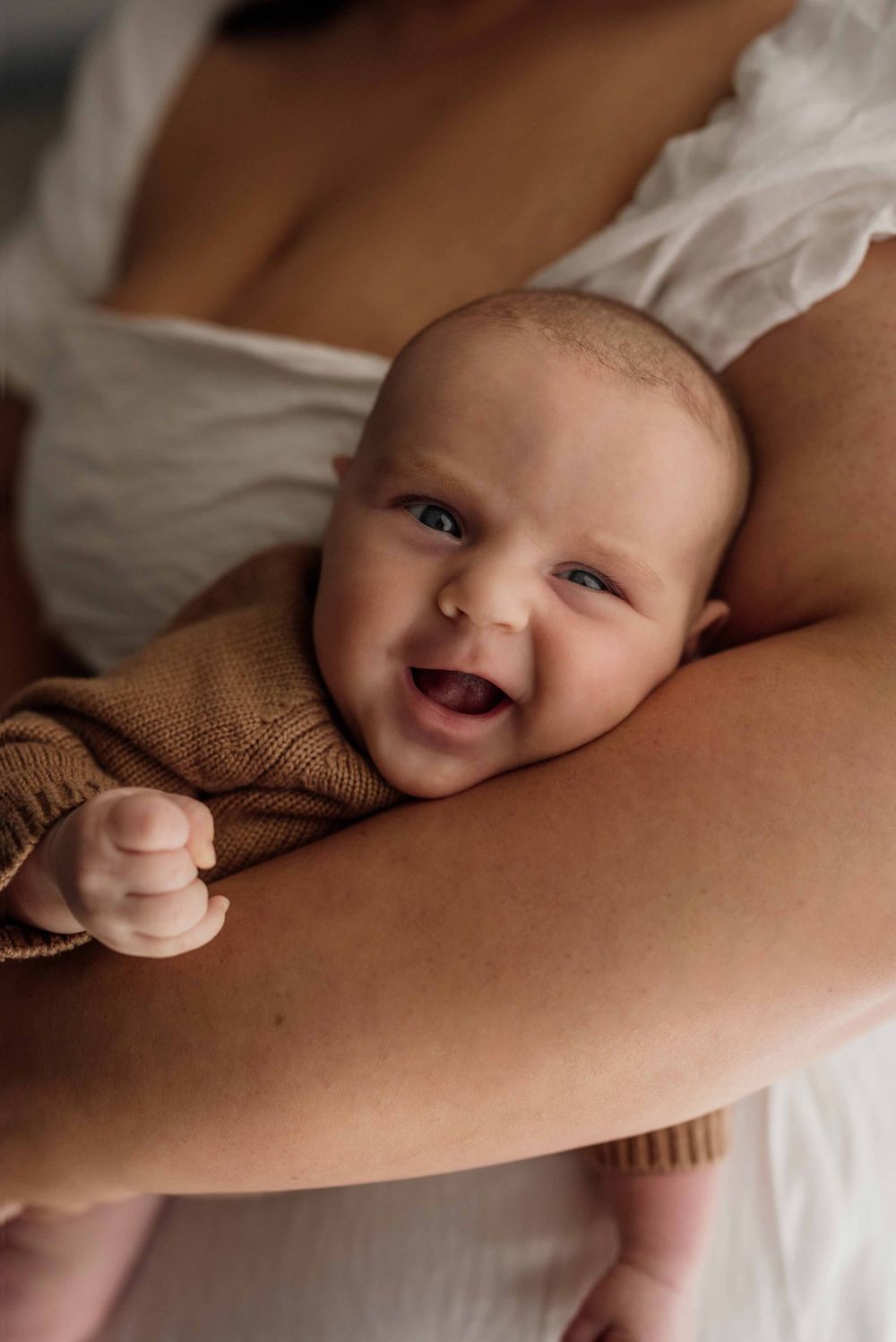 Smiling newborn baby in brown knit outfit cradled in mother's arms during newborn photography session.