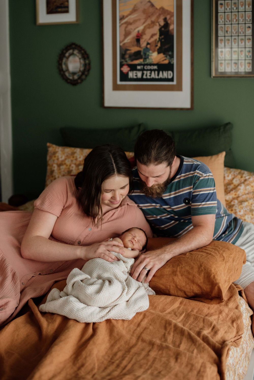 Parents smiling at swaddled newborn on bed with green walls and New Zealand travel poster decor.