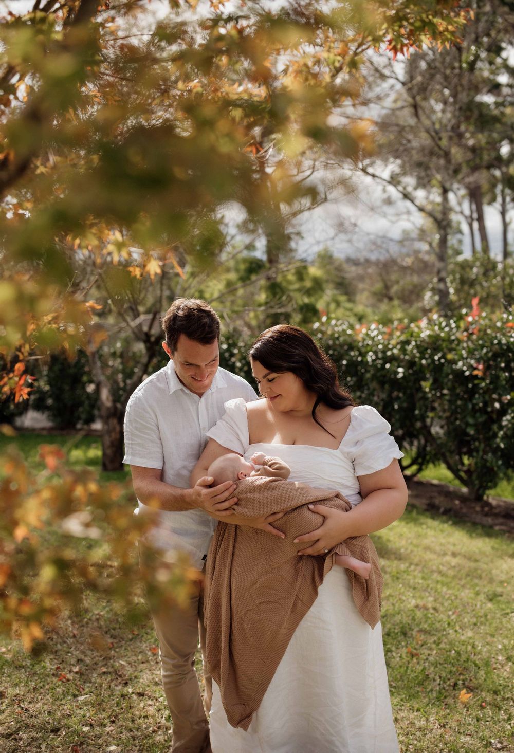 Couple standing in autumn garden tenderly looking at newborn baby wrapped in tan muslin cloth.