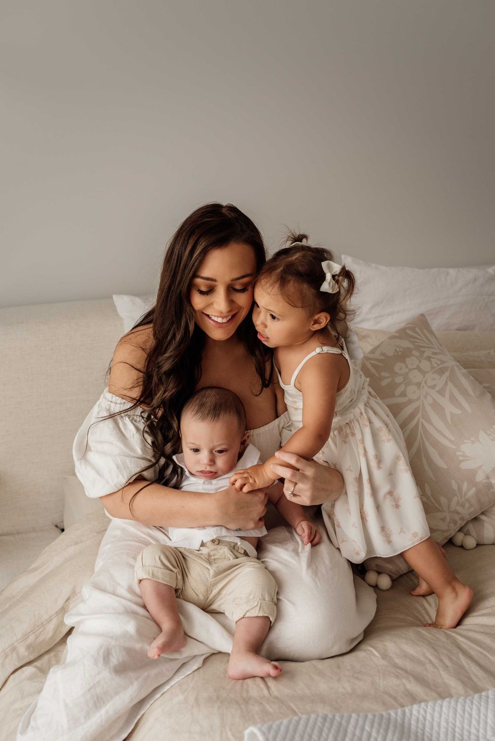 Mother sitting on bed smiling at baby boy while toddler daughter in white dress leans in close.