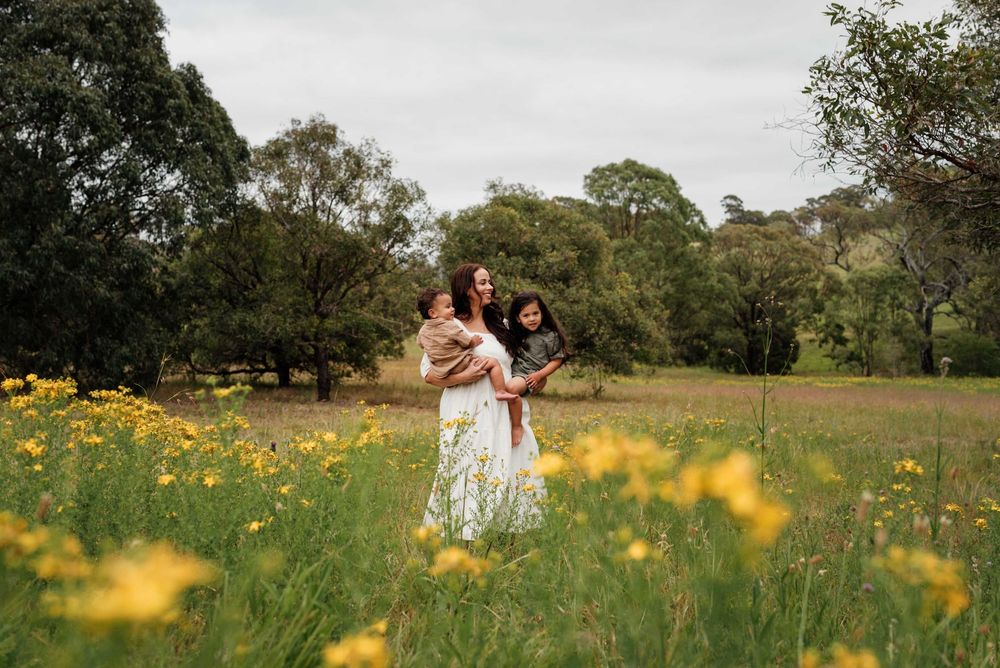 Mother in white dress holding two children in yellow wildflower meadow.