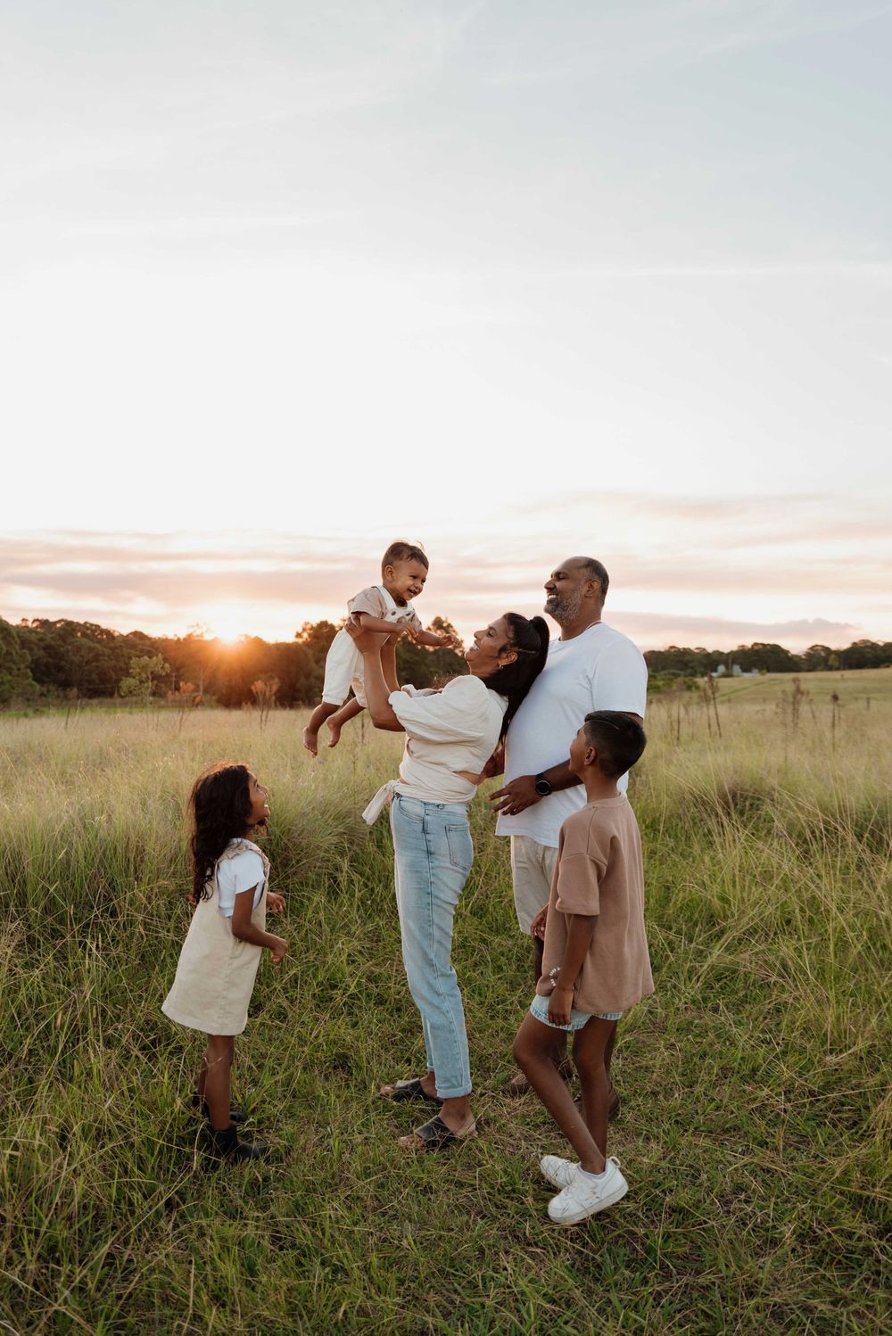 Family of five enjoying sunset in open grassy field, mother lifting baby.