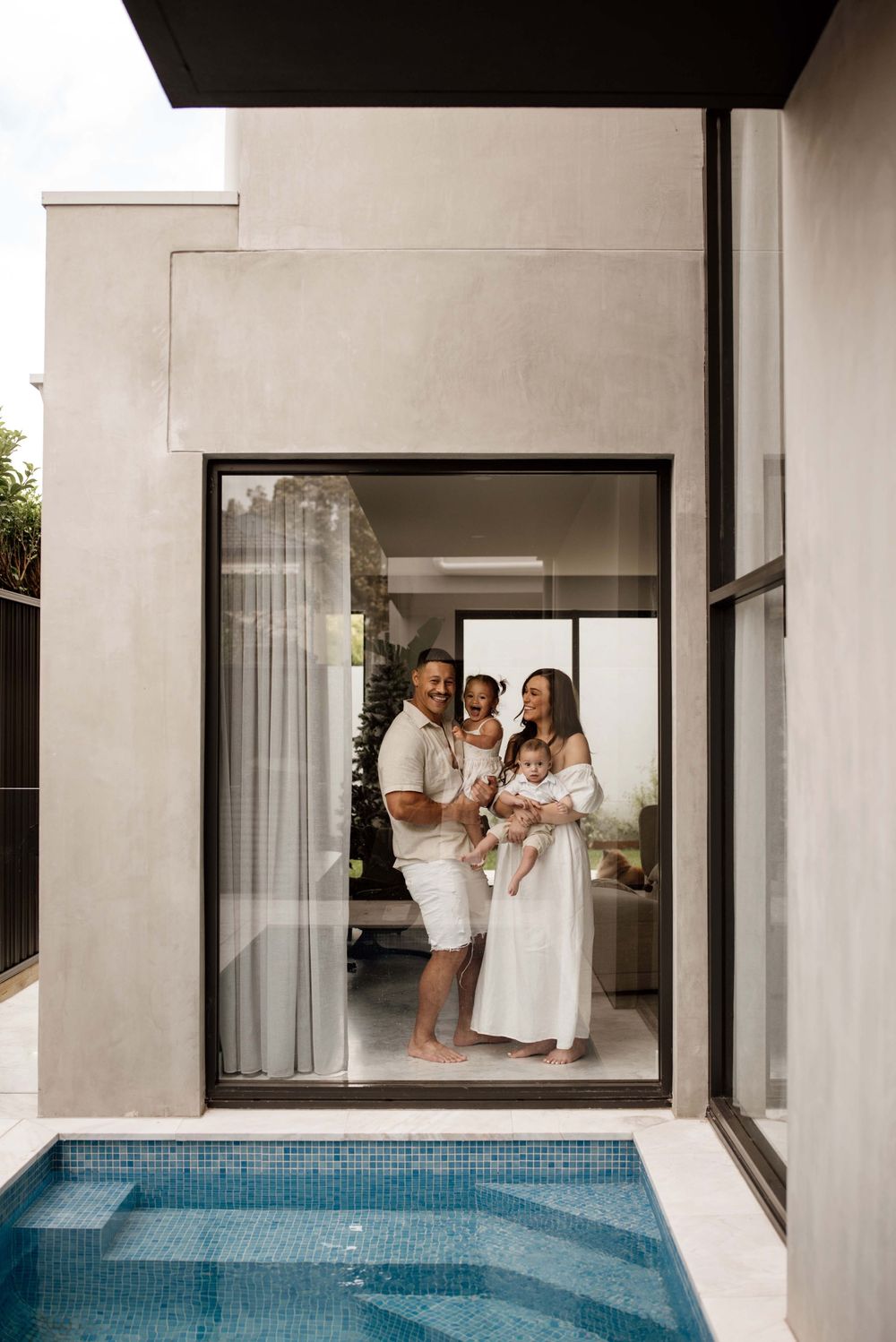 Family of four in white outfits posing in large glass doorway beside pool.