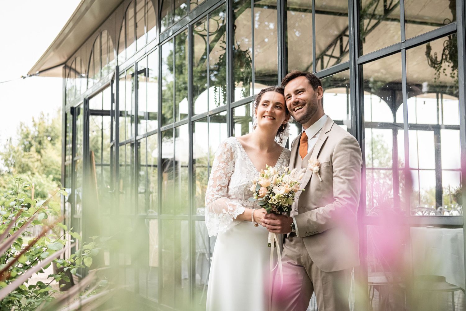 Elegant couple poses in sunlit greenhouse with vintage architecture and soft natural lighting.