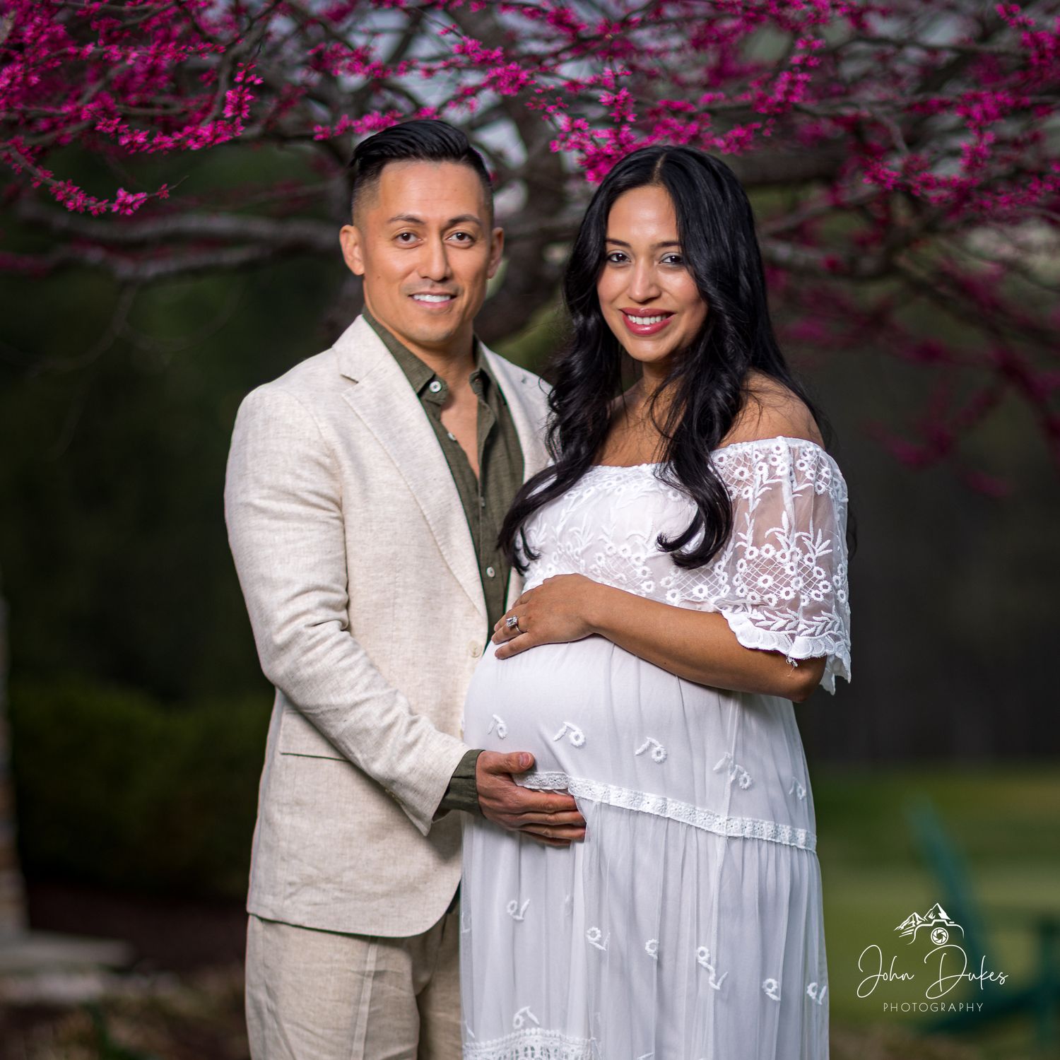 An expecting couple poses together under blooming cherry blossoms during maternity photoshoot.