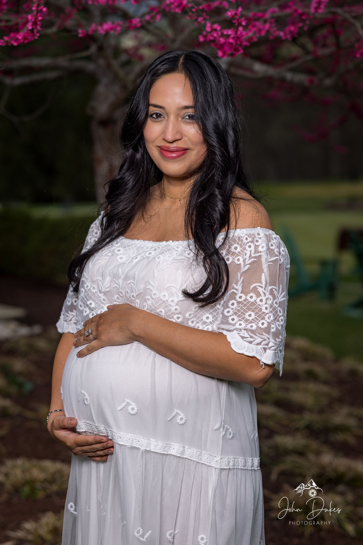 A woman in white lace dress poses for maternity photos with blooming cherry blossoms in background.